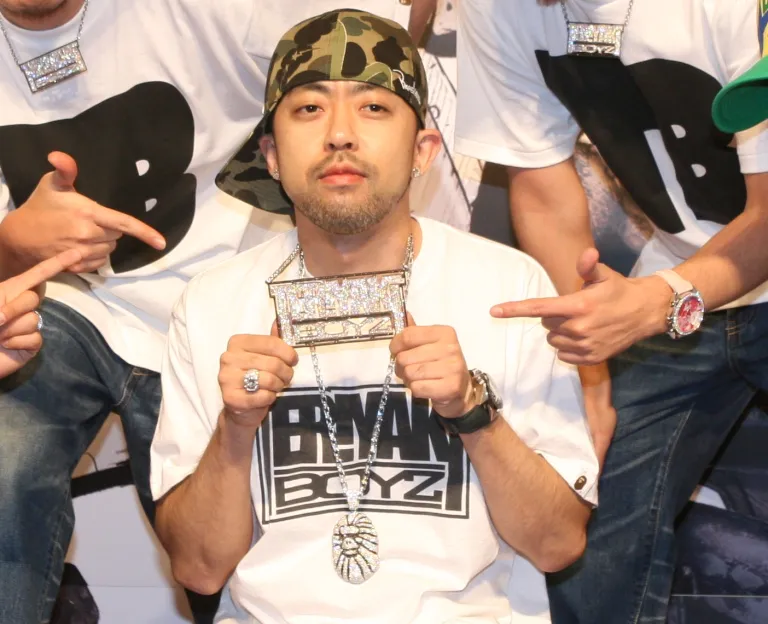 NIGO wearing a camouflage bucket hat and a 'Billionaire Boyz' graphic T-shirt, holding up a large diamond-encrusted 'Billionaire Boyz' chain necklace, surrounded by others pointing at it