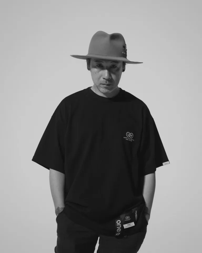 Undercover founder Jun Takahashi wearing a wide-brim felt hat and an oversized black Undercover x WTAPS collab T-shirt, photographed in black and white against a plain background