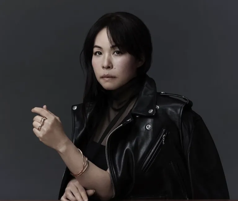Sacai founder Chitose Abe wearing a black leather biker jacket, photographed against a dark grey background, with rose gold rings and bangles on her hand