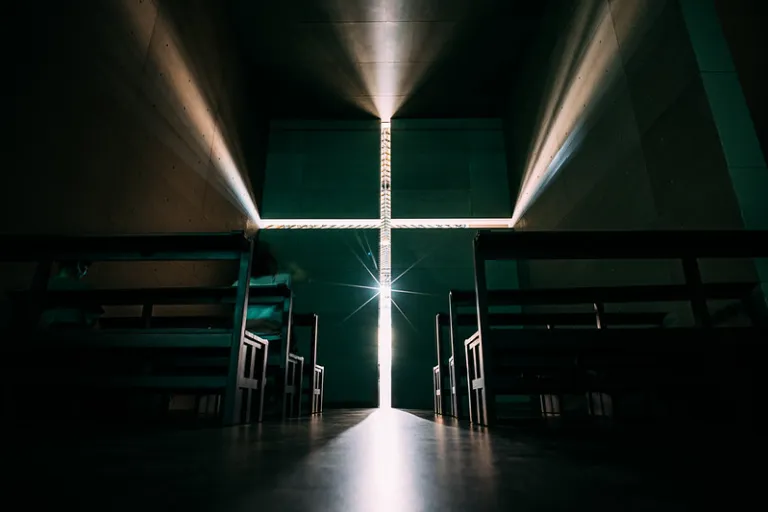 Interior of the Church of the Light, with sunlight forming a bright cross on the concrete wall between dark wooden pews.