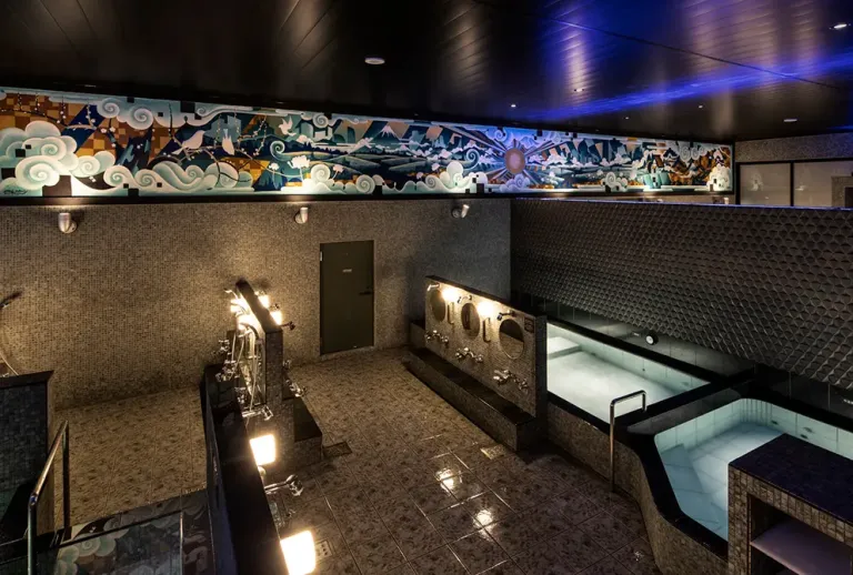 Interior of Kairyou-yu, featuring dark tiled baths, washing stations and a colourful mural above the bathing area.