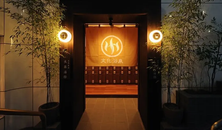 Entrance of Bunkayokusen, a stylish sento in Tokyo, warmly lit with bamboo plants and a noren curtain.