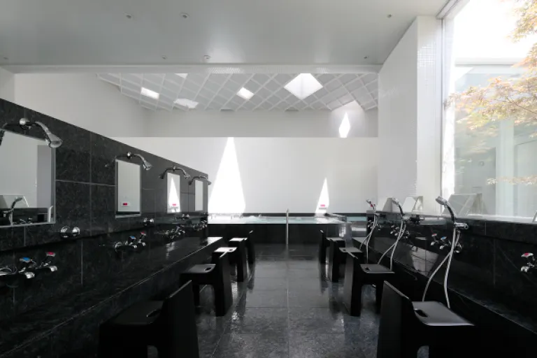 Bright washing area inside Hisamatsuyu, featuring black tiled walls, low stools and natural light from a large window.