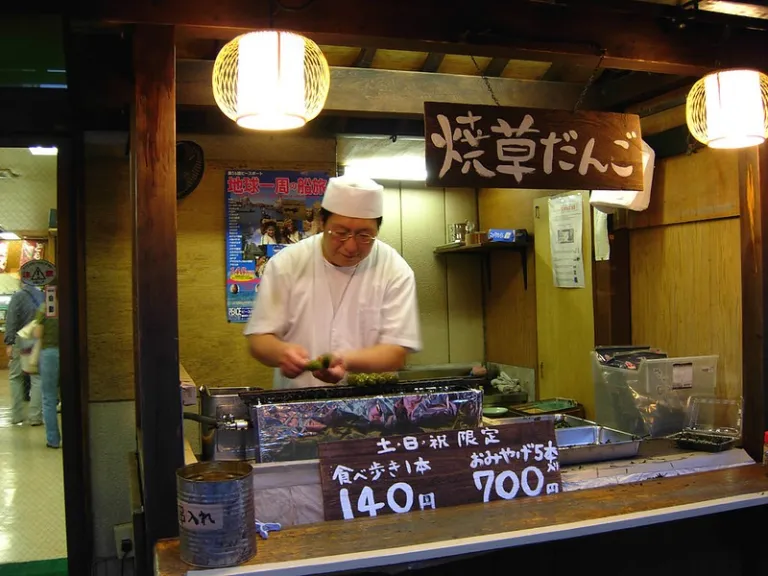 Staff member grilling kusa dango at a traditional shop in Shibamata, with a sign advertising the local speciality.