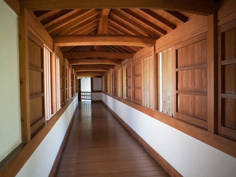 Interior view of a long wooden corridor with a traditional Japanese post-and-beam ceiling and shoji screen windows.