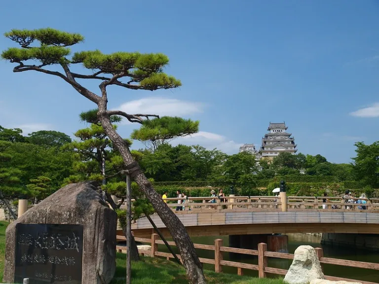 A scenic view of a stylized pine tree, an inscribed stone monument, and a wooden bridge leading towards Himeji Castle under a clear blue sky