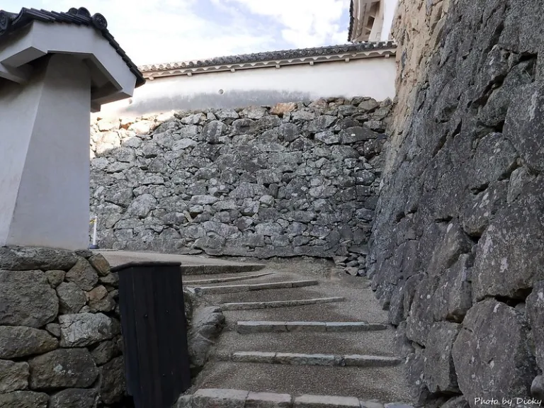A narrow stone staircase curving upward, flanked by massive stone walls and a traditional plaster wall at Himeji Castle.