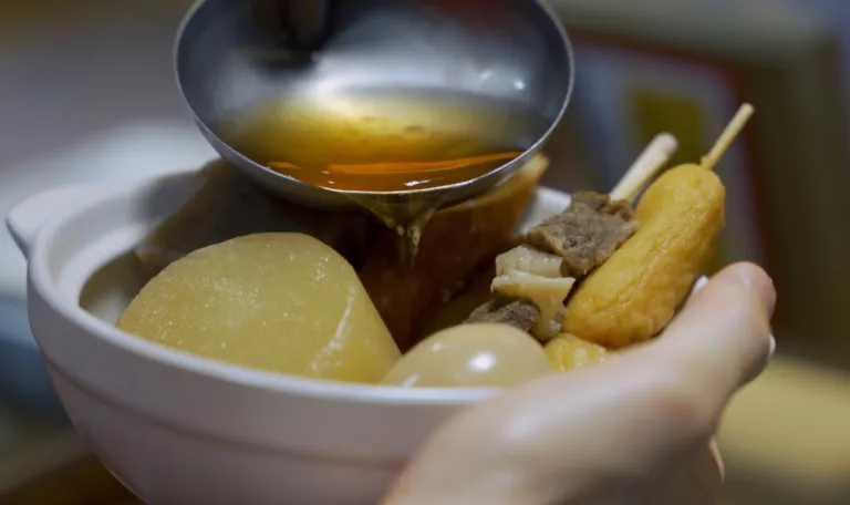 Clear dashi broth being poured from a ladle into a bowl of Japanese oden with radish, boiled egg, and skewers.