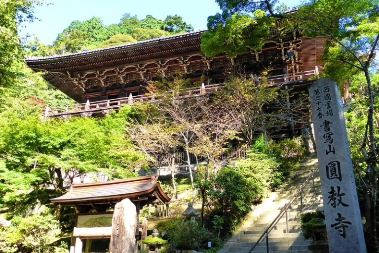 Stone steps leading up to Manidono, a historic wooden temple hall built on stilt pillars, surrounded by lush green trees at Shoshazan Engyo-ji Temple.