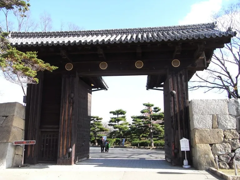 A massive dark-wooden traditional gate with tiled roof and gold crests, opening into a garden area at Himeji Castle.